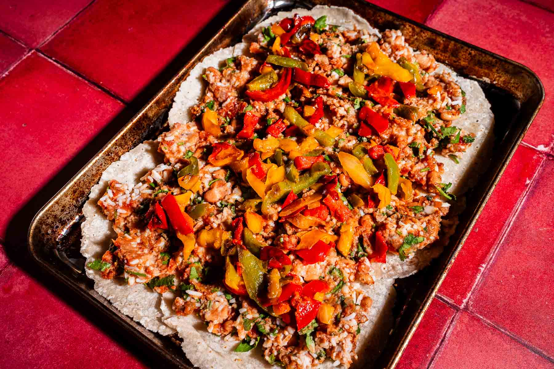 A baking tray on a red tiled surface holds a flat dish, reminiscent of a sheet pan crunchwrap, topped with rice, beans, and sliced red, yellow, and green peppers.