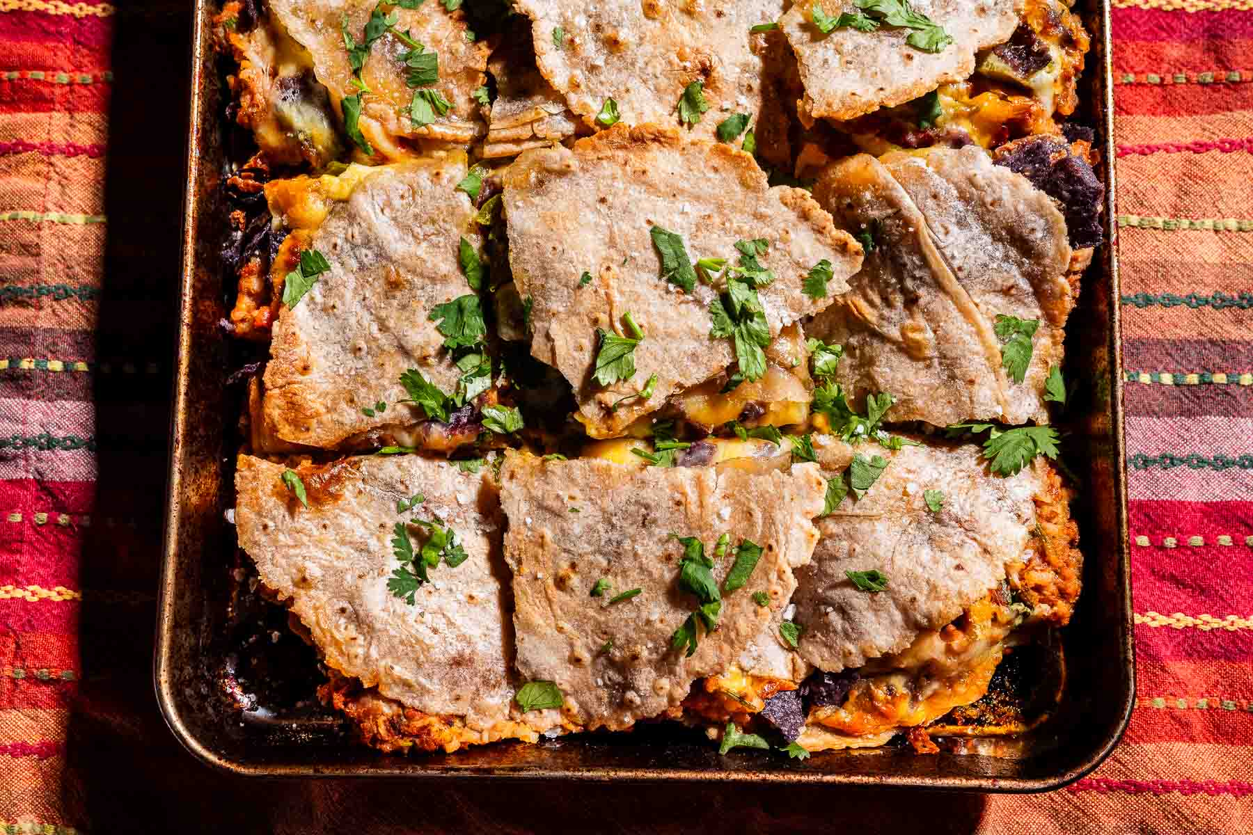 A baking dish with square slices of a layered sheet pan crunchwrap casserole topped with chopped cilantro, on a colorful woven tablecloth.