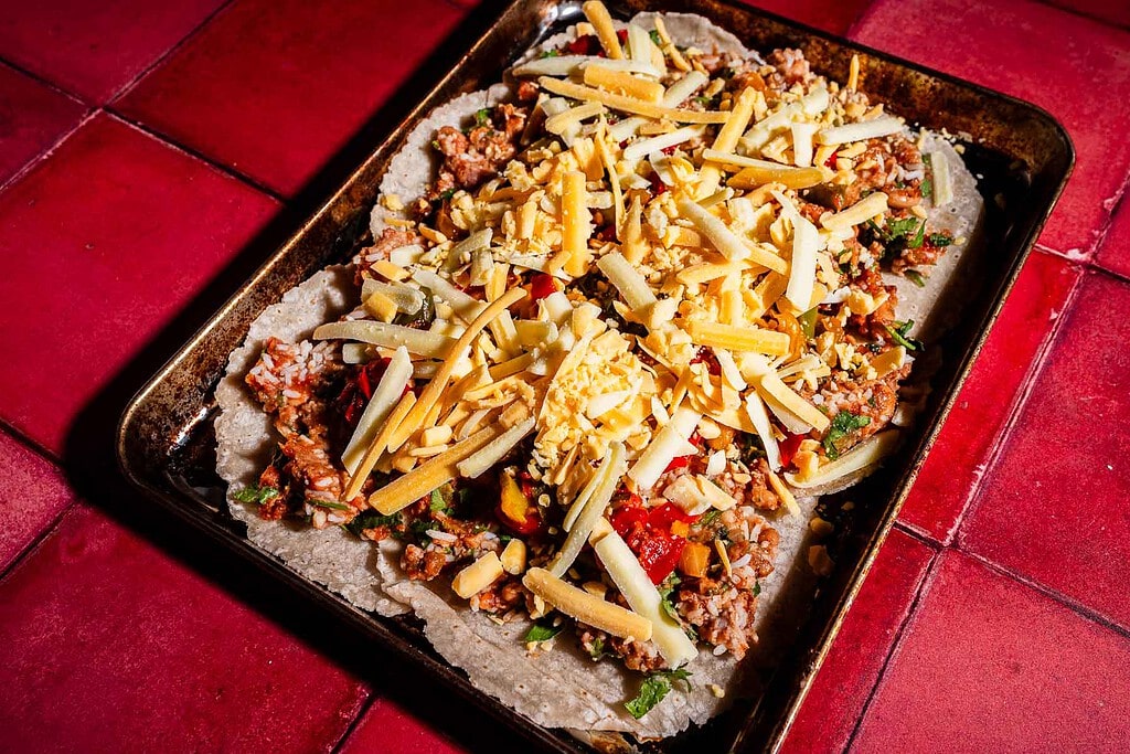 A baking tray on a red tiled surface holds an uncooked sheet pan crunchwrap, topped with shredded cheese, ground meat, and vegetables on a flat dough base.