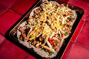 A baking tray on a red tiled surface holds an uncooked sheet pan crunchwrap, topped with shredded cheese, ground meat, and vegetables on a flat dough base.