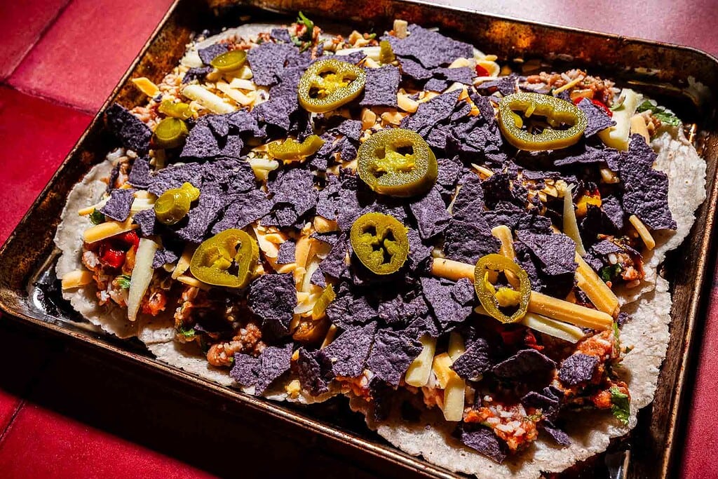 A baking sheet covered with nachos topped with blue corn tortilla chips, shredded cheese, ground meat, vegetables, and sliced jalapeños for a sheet pan crunchwrap-inspired twist.