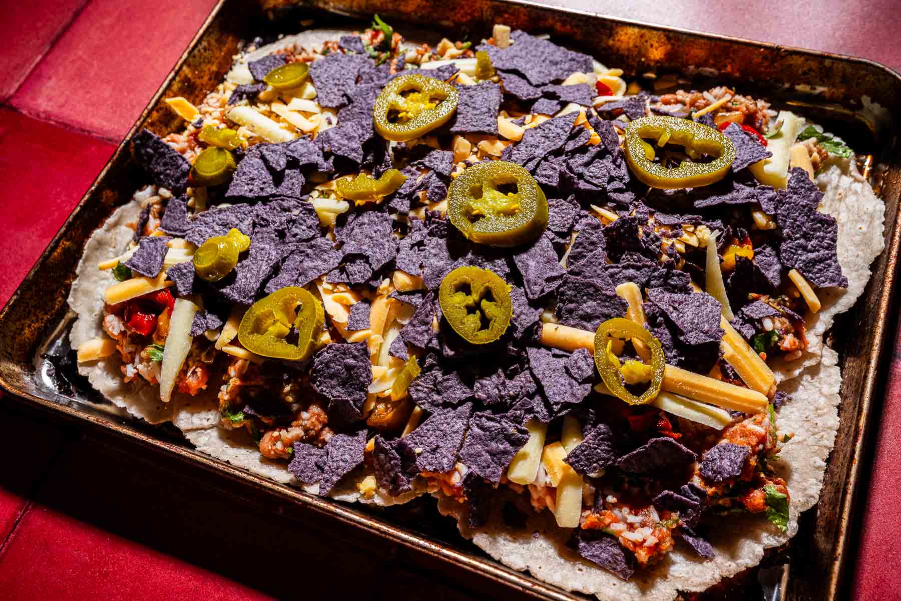 A baking sheet covered with nachos topped with blue corn tortilla chips, shredded cheese, ground meat, vegetables, and sliced jalapeños for a sheet pan crunchwrap-inspired twist.