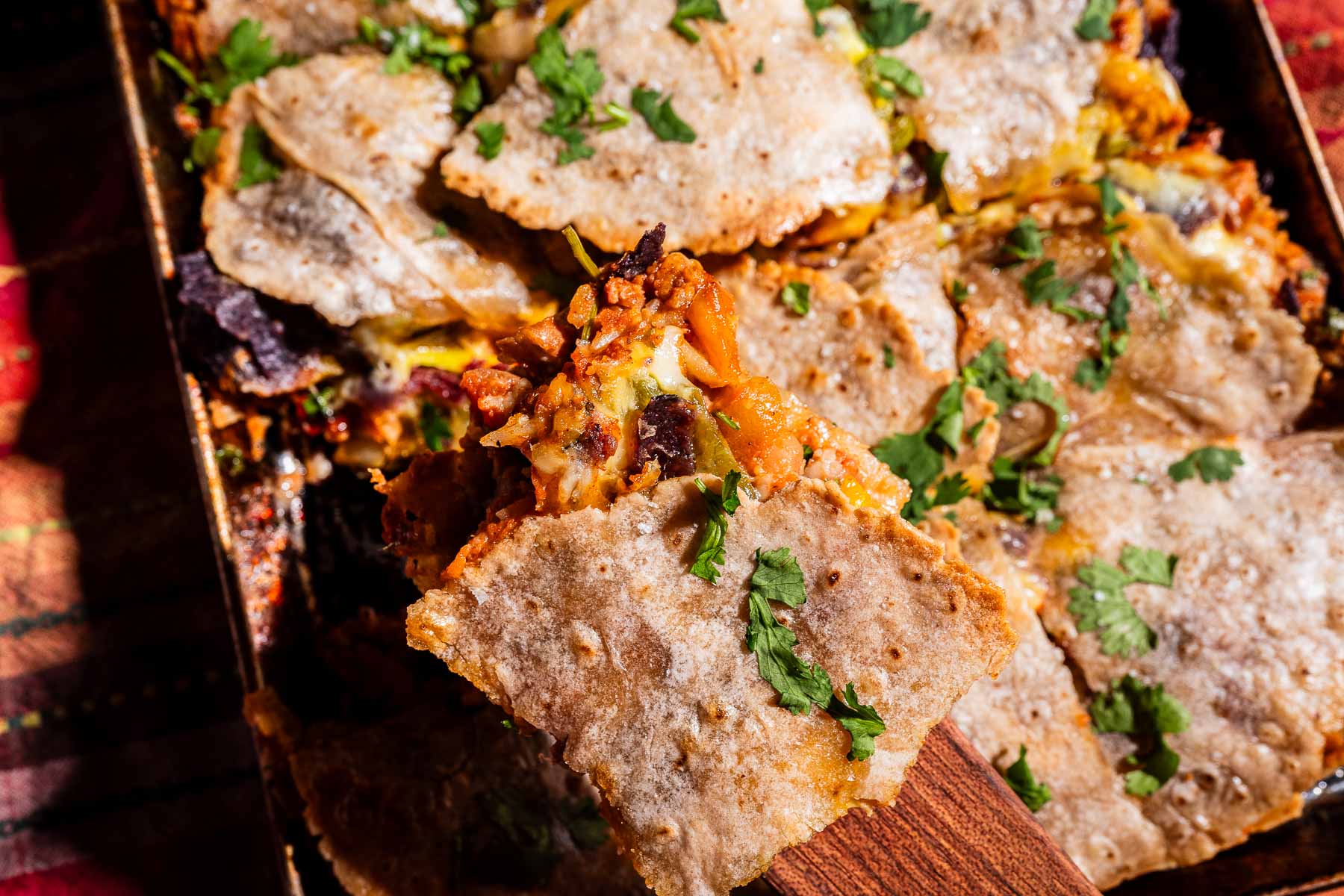 A spatula lifts a serving of sheet pan crunchwrap-style baked quesadilla casserole, topped with melted cheese and fresh cilantro, from a tray.