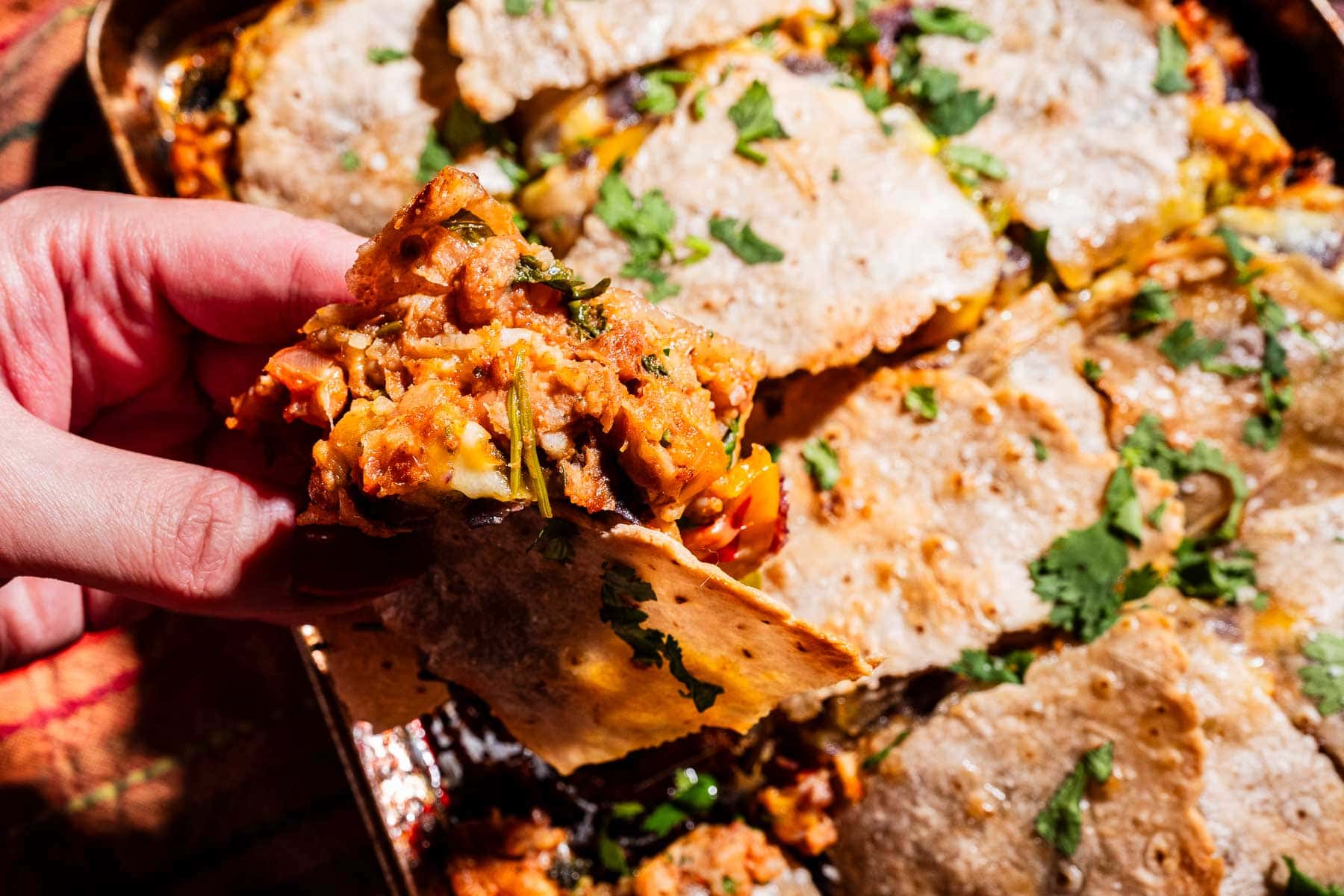 A hand holding a slice of baked casserole with a crispy topping, filled with vegetables and garnished with chopped cilantro, reminiscent of a sheet pan crunchwrap.