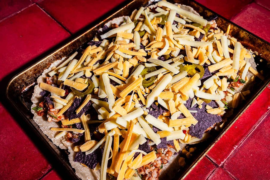 A baking sheet holds tortilla chips topped with ground meat, jalapeños, and shredded cheese, creating a sheet pan crunchwrap vibe on a red tiled surface.
