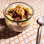 A glass jar filled with banana chia pudding, granola, and banana slices sits on a tiled surface next to a metal spoon.