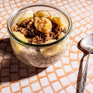 A glass jar filled with banana chia pudding, granola, and banana slices sits on a tiled surface next to a metal spoon.