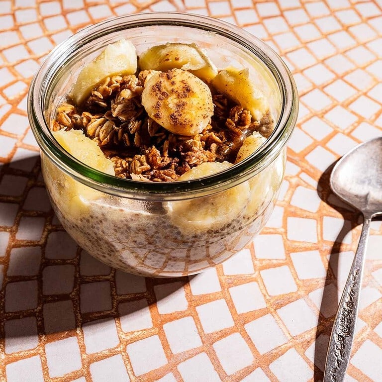 A glass jar filled with banana chia pudding, granola, and banana slices sits on a tiled surface next to a metal spoon.