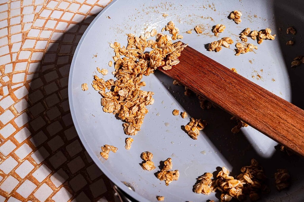 A close-up of oats and a wooden spatula in a lightly used frying pan on a tiled surface, evoking the cozy preparation of banana chia pudding, with light casting gentle shadows across the scene.