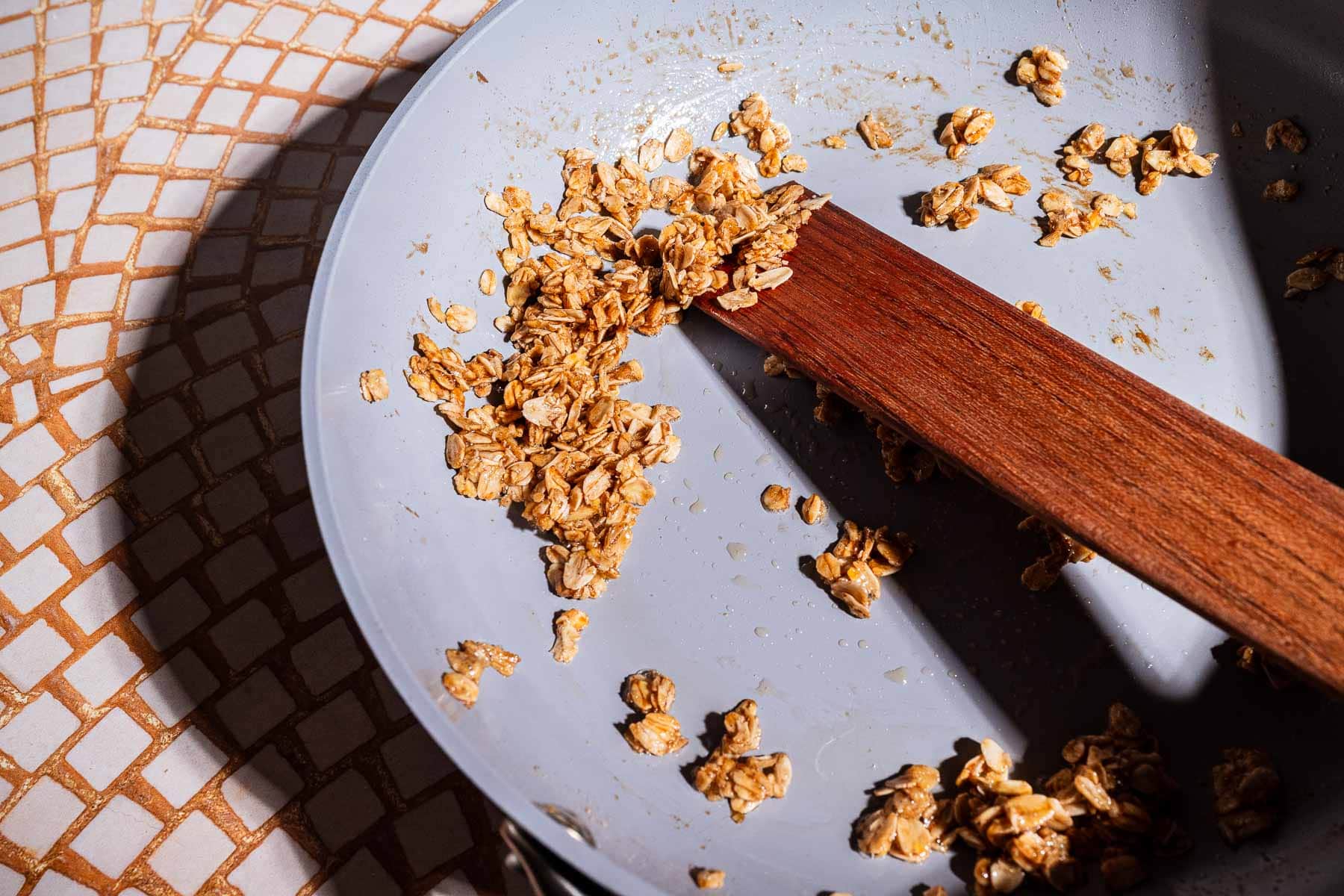 A close-up of oats and a wooden spatula in a lightly used frying pan on a tiled surface, evoking the cozy preparation of banana chia pudding, with light casting gentle shadows across the scene.