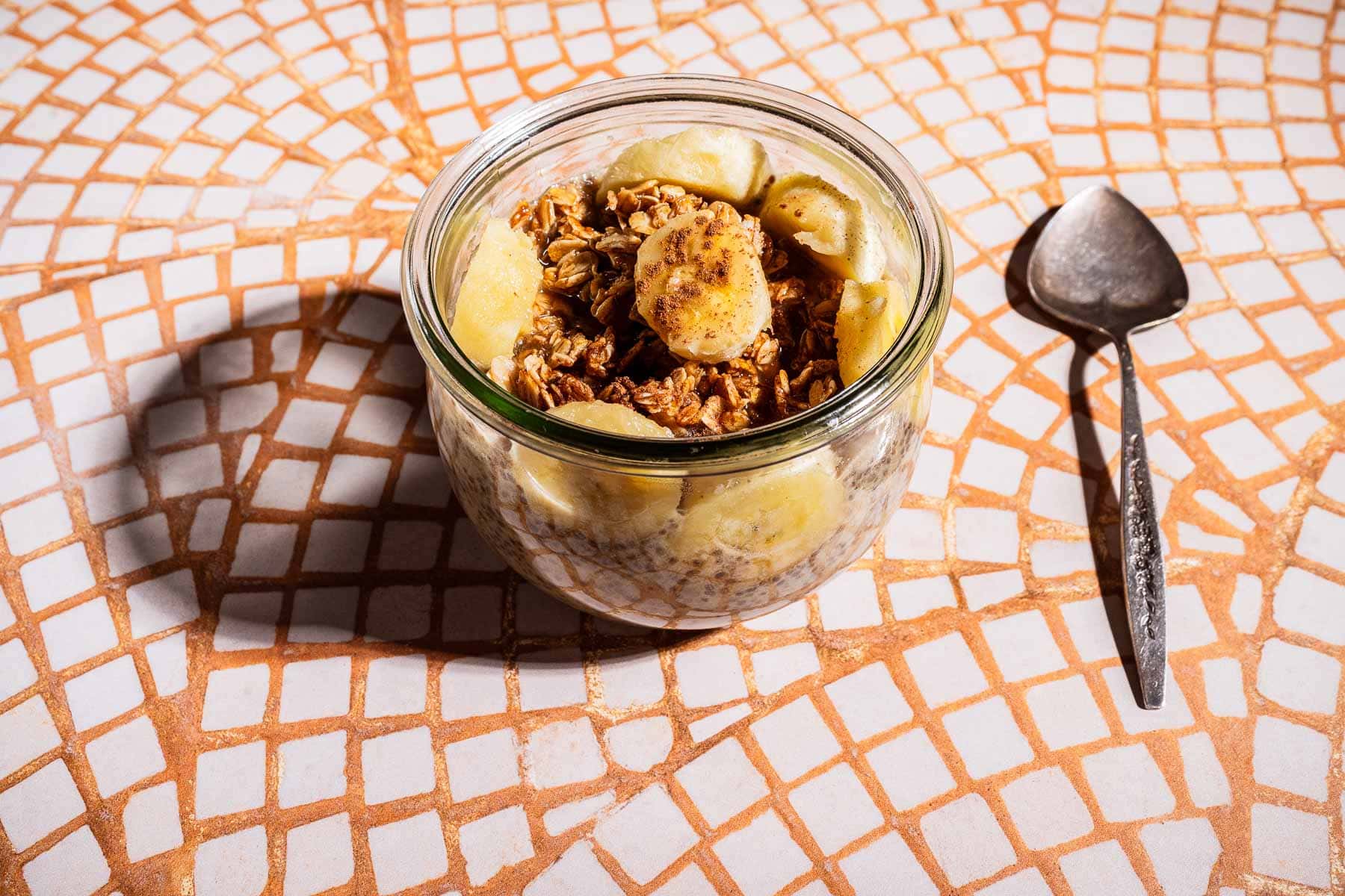A glass jar filled with granola, oats, and banana chia pudding sits on a patterned tile surface next to a spoon.
