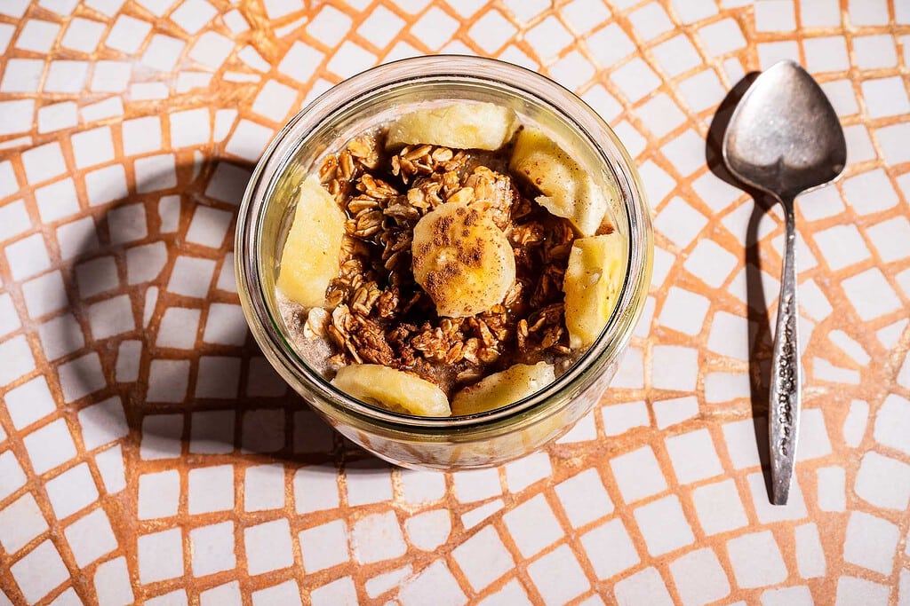 A jar filled with banana chia pudding, topped with granola and banana slices, sits on a mosaic-tiled surface next to a metal spoon.