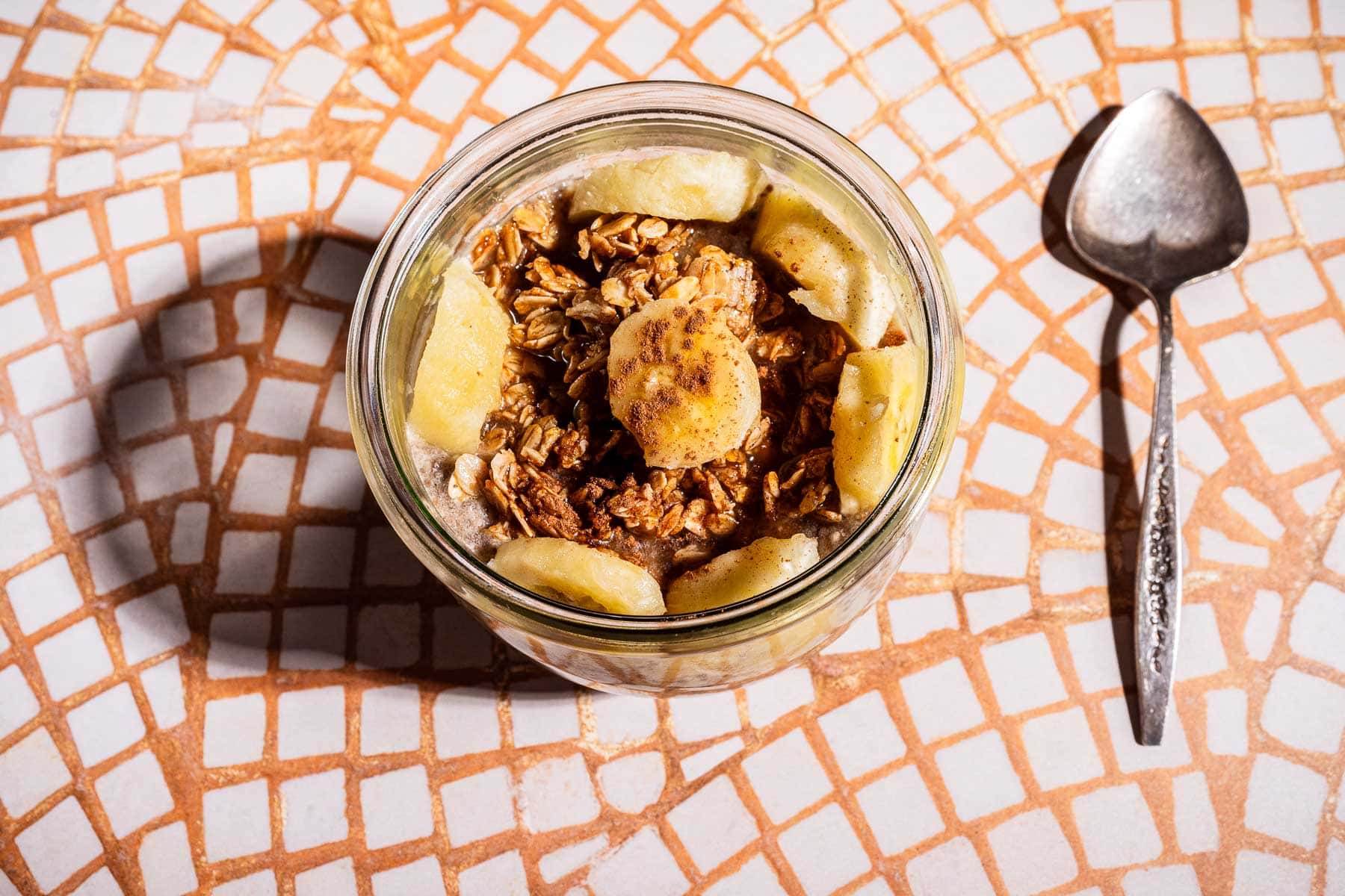 A jar filled with banana chia pudding, topped with granola and banana slices, sits on a mosaic-tiled surface next to a metal spoon.