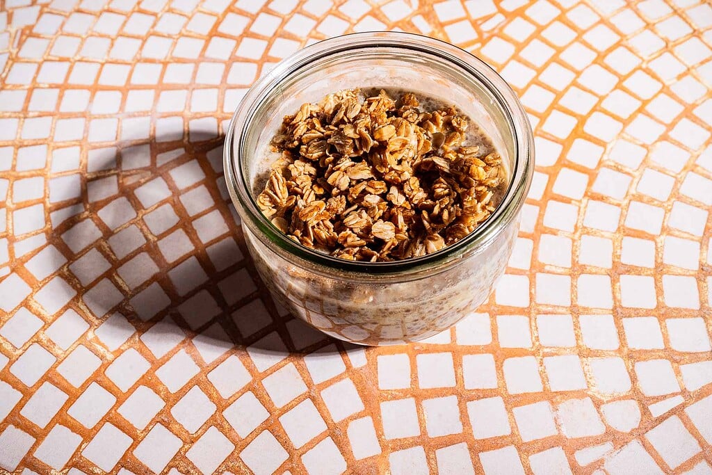 A glass jar filled with overnight oats and banana chia pudding, topped with granola, sits on a tile-patterned surface.
