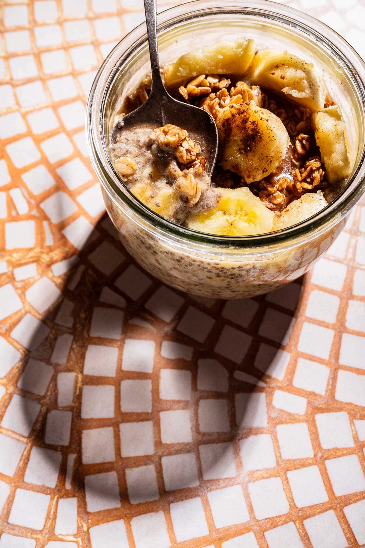 A glass jar filled with banana chia pudding, oatmeal, and granola sits on a patterned surface with a spoon inside.