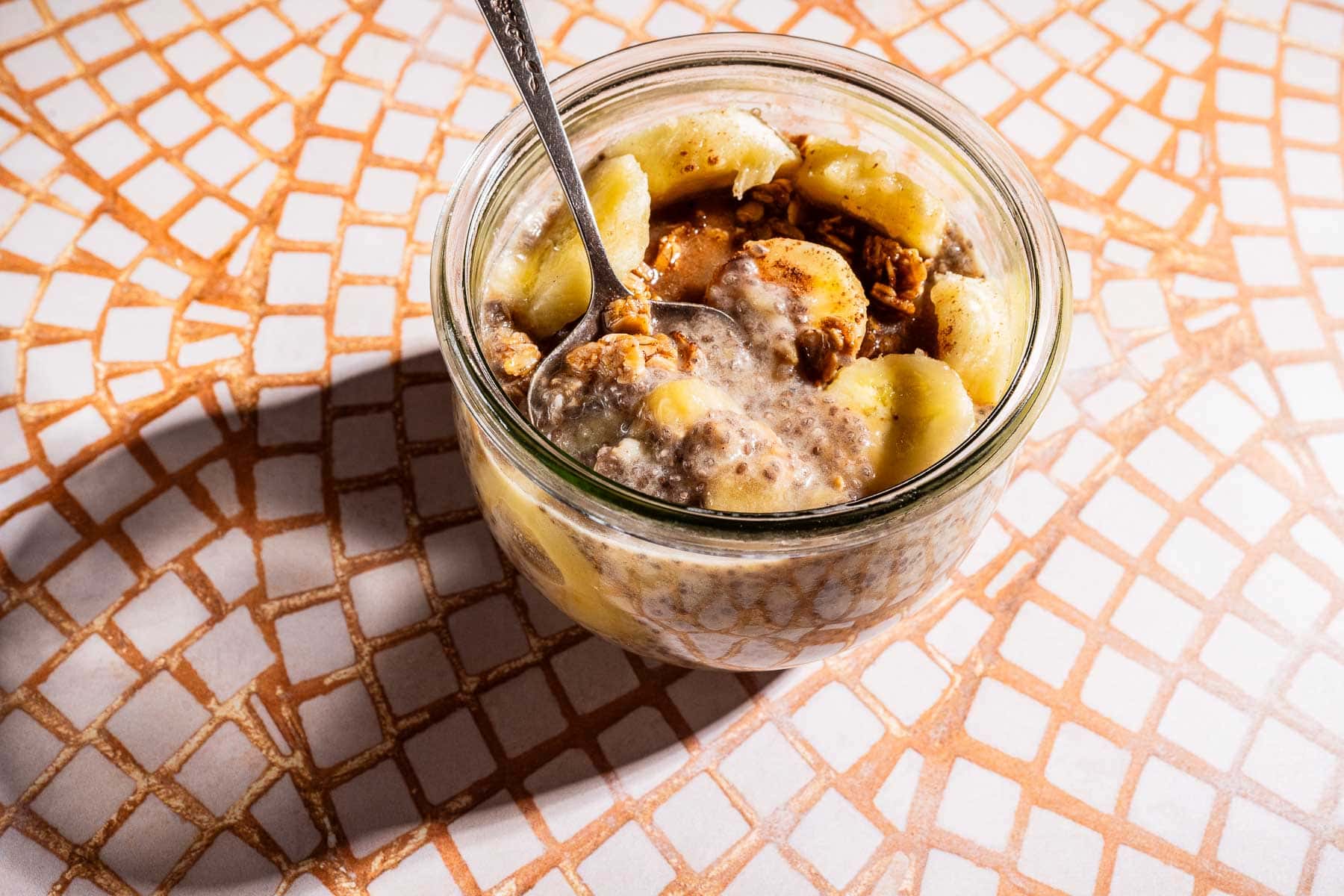 A glass jar filled with banana chia pudding, topped with sliced bananas and granola, with a spoon inside, sits on a mosaic tile surface.