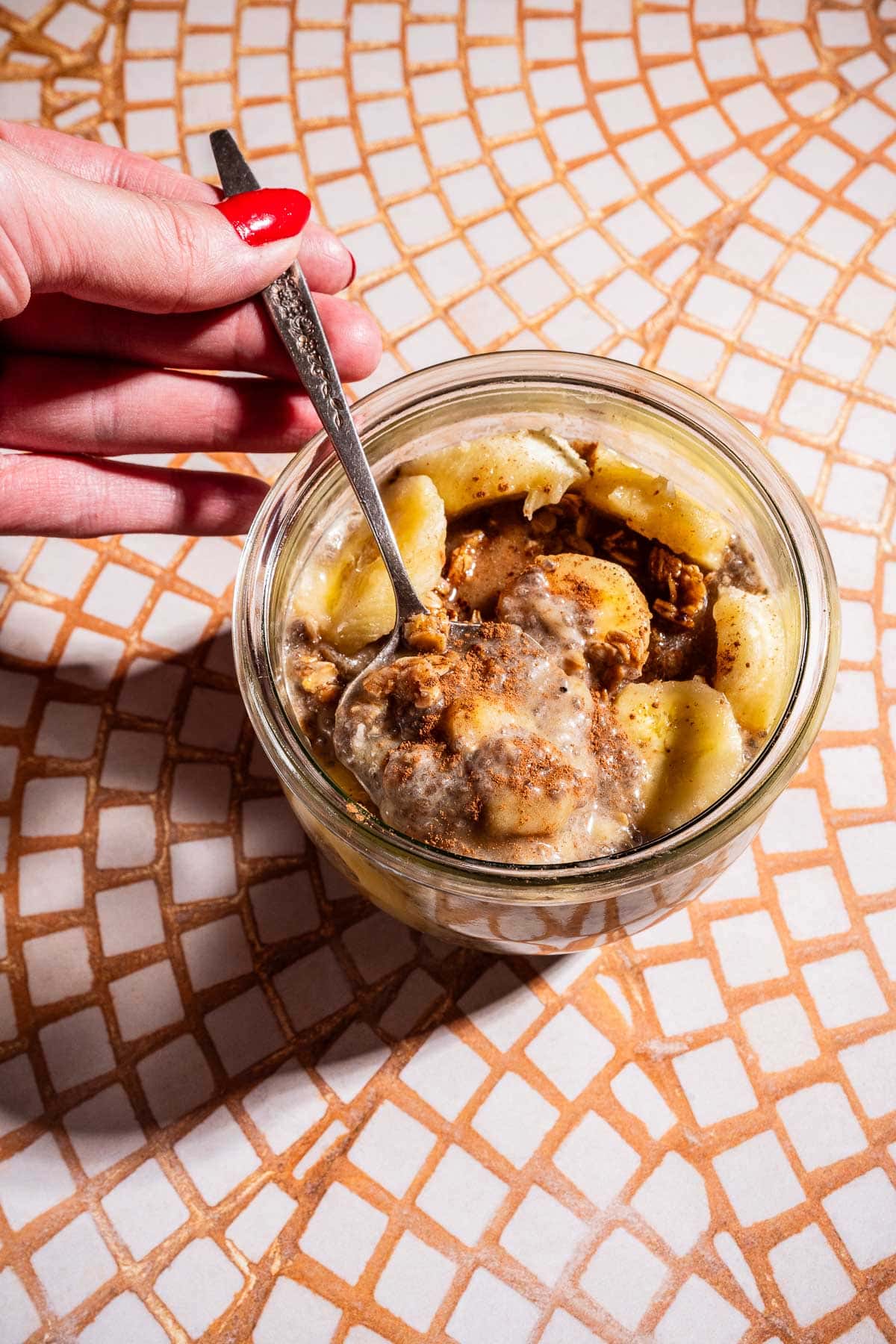 A hand holding a spoon over a glass jar filled with sliced bananas, oatmeal, nut butter, and creamy banana chia pudding on a mosaic-tiled surface.