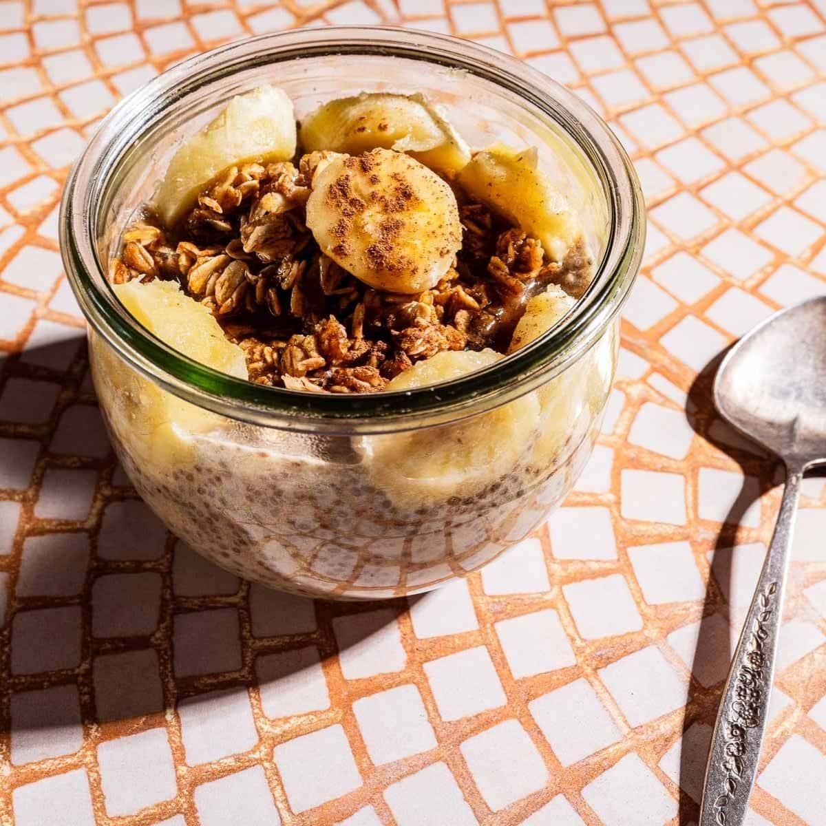 A glass jar filled with banana chia pudding, granola, and banana slices sits on a tiled surface next to a metal spoon.