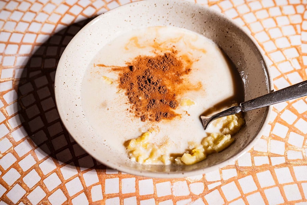 A bowl with mashed banana, milk, and a heap of ground cinnamon—perfect for making banana chia pudding—rests on a tiled surface with a spoon inside.