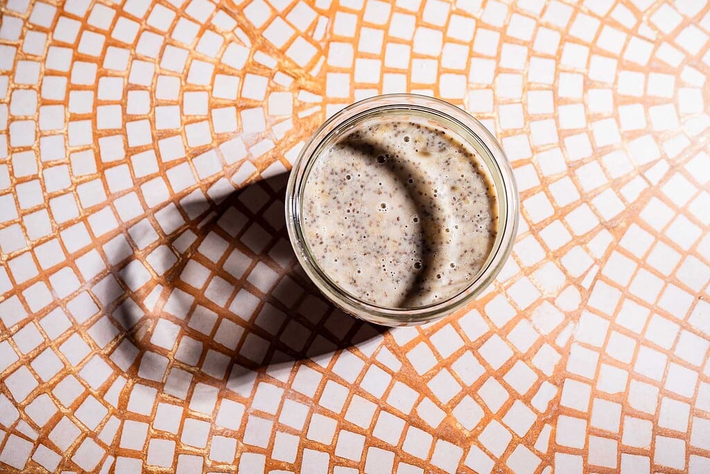 A glass jar filled with a speckled, creamy banana chia pudding smoothie sits on a patterned tile surface, casting a shadow to the left.