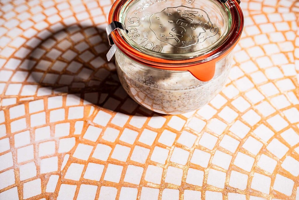 A glass jar with a metal clip and orange seal contains banana chia pudding, placed on a patterned tile surface with a shadow to the left.