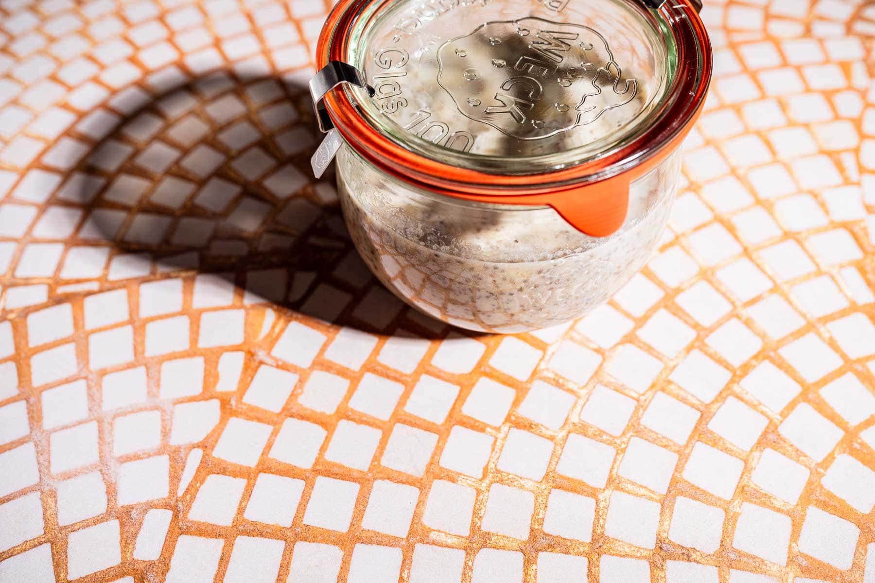 A glass jar with a metal clip and orange seal contains banana chia pudding, placed on a patterned tile surface with a shadow to the left.