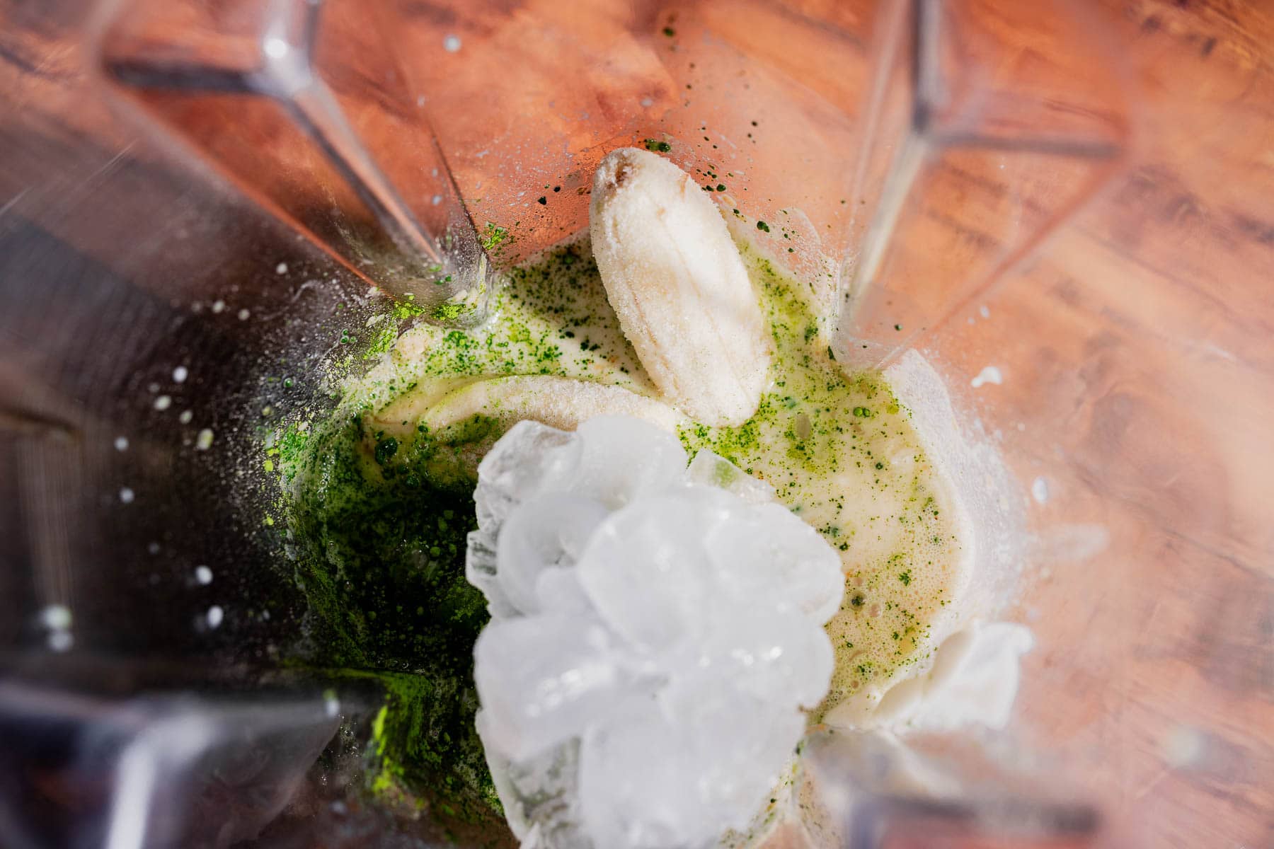 A blender contains banana, ice, green powder, and liquid on a wooden surface, viewed from above—perfect for making a refreshing matcha protein shake.
