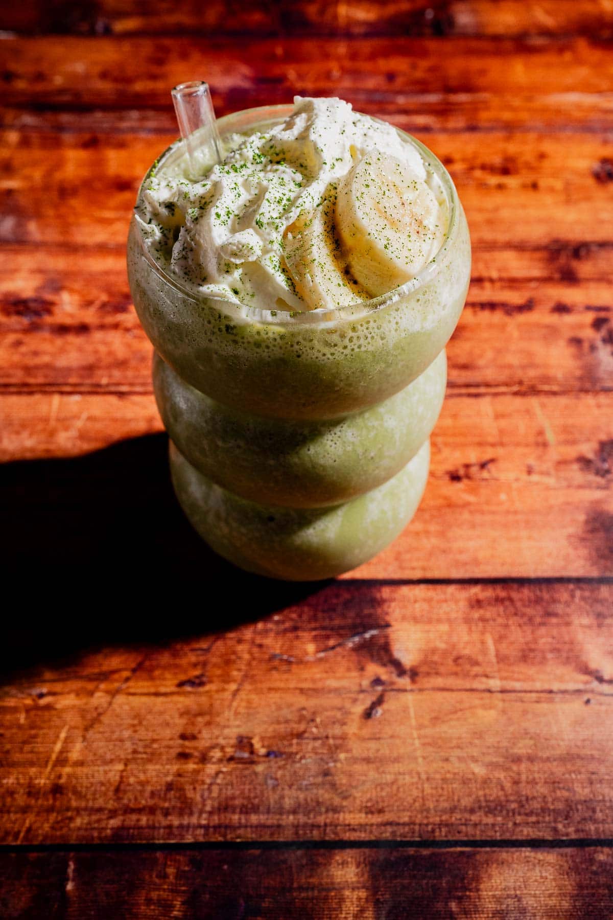 A matcha protein shake in a round glass, topped with whipped cream, banana slices, and green powder, sits on a wooden surface.