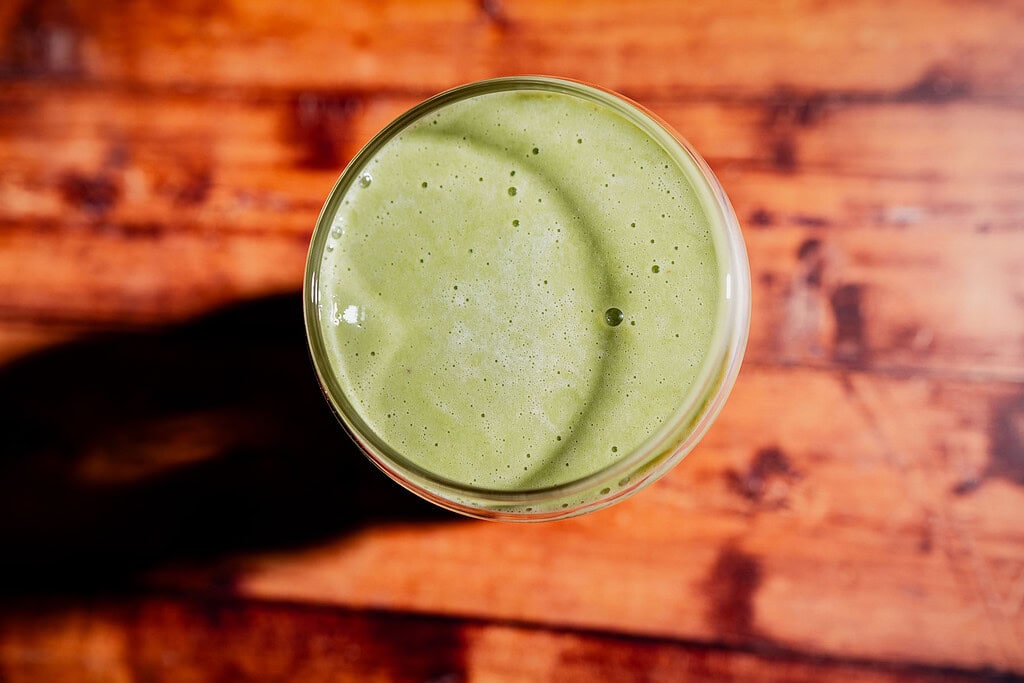 A glass of matcha protein shake viewed from above, placed on a wooden surface with visible shadows.