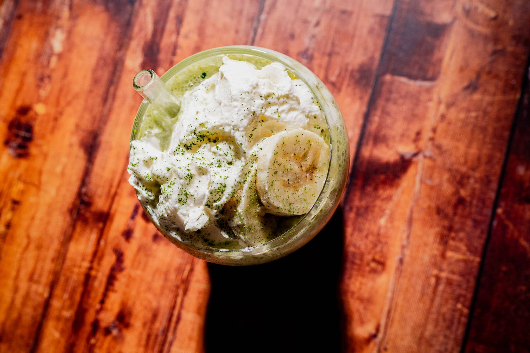 A glass of matcha protein shake topped with whipped cream, banana slices, and a sprinkle of green powder, placed on a wooden surface.