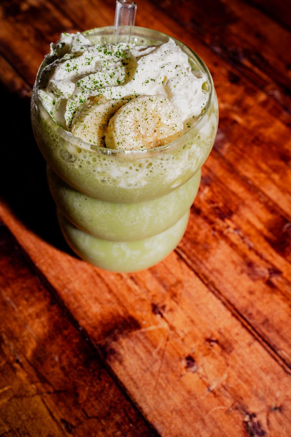A matcha protein shake topped with whipped cream, banana slices, and a sprinkle of green powder, served in a curvy glass on a wooden table.
