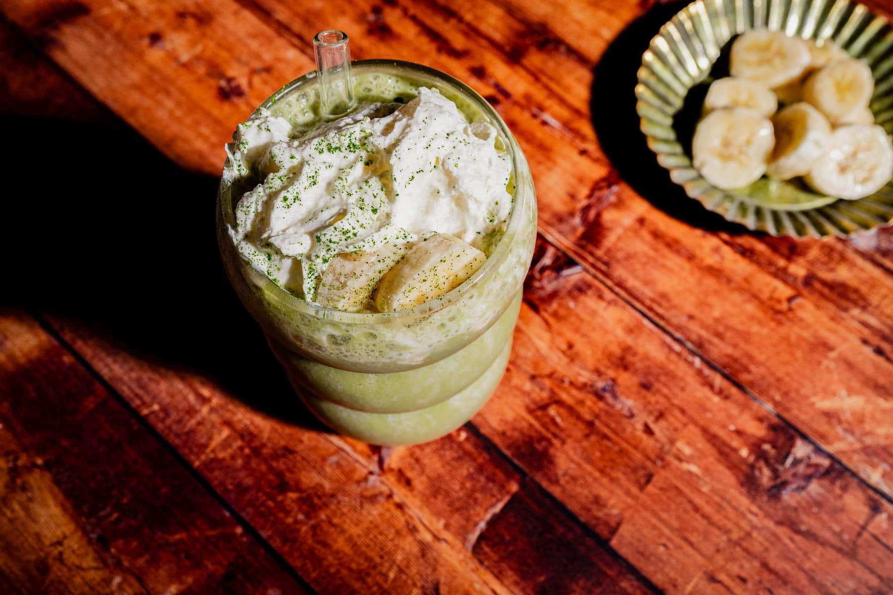 A glass of matcha protein shake topped with whipped cream and banana slices sits on a wooden table, with a small plate of sliced bananas in the background.