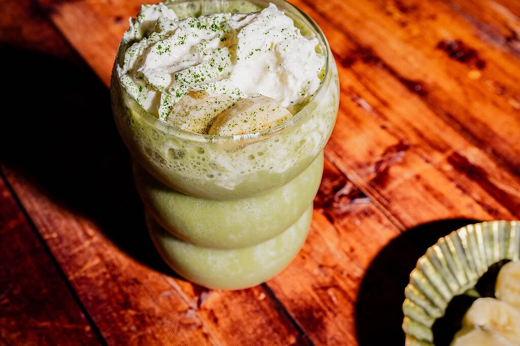 A green smoothie in a round glass topped with whipped cream, banana slices, and green powder sits on a wooden surface—enjoy this vibrant matcha protein shake for a delicious boost.