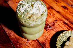 A green smoothie in a round glass topped with whipped cream, banana slices, and green powder sits on a wooden surface—enjoy this vibrant matcha protein shake for a delicious boost.