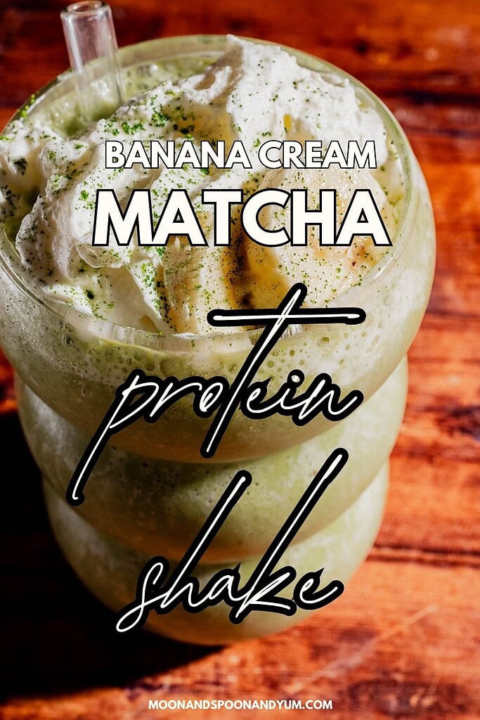 A glass of matcha protein shake with banana cream, topped with whipped cream and set on a wooden surface. Text overlay reads "Banana Cream Matcha Protein Shake.