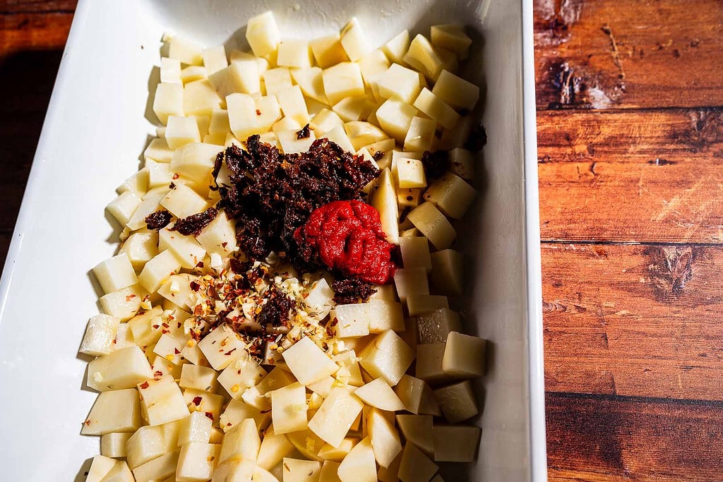Diced potatoes in a white dish topped with chopped sun-dried tomatoes, tomato paste, minced garlic, and red pepper flakes—a delicious spin on marry me potatoes—served on a wooden surface.