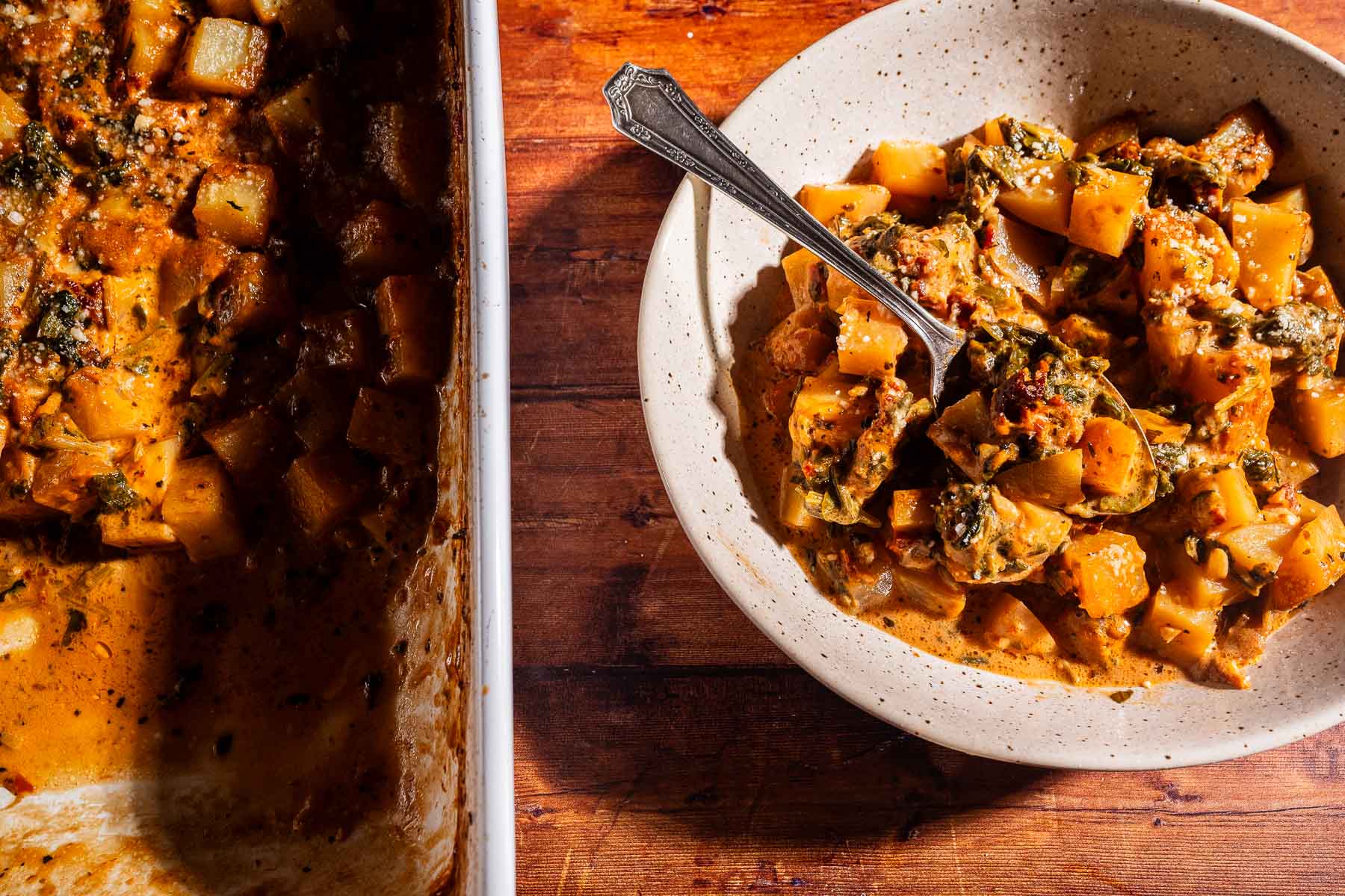A bowl of baked vegetable stew with a spoon, next to a casserole dish containing the remaining stew and some hearty marry me potatoes, all set on a wooden table.