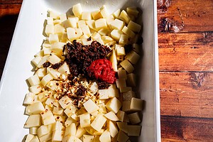 Diced potatoes in a white dish topped with chopped sun-dried tomatoes, tomato paste, minced garlic, and red pepper flakes—a delicious spin on marry me potatoes—served on a wooden surface.