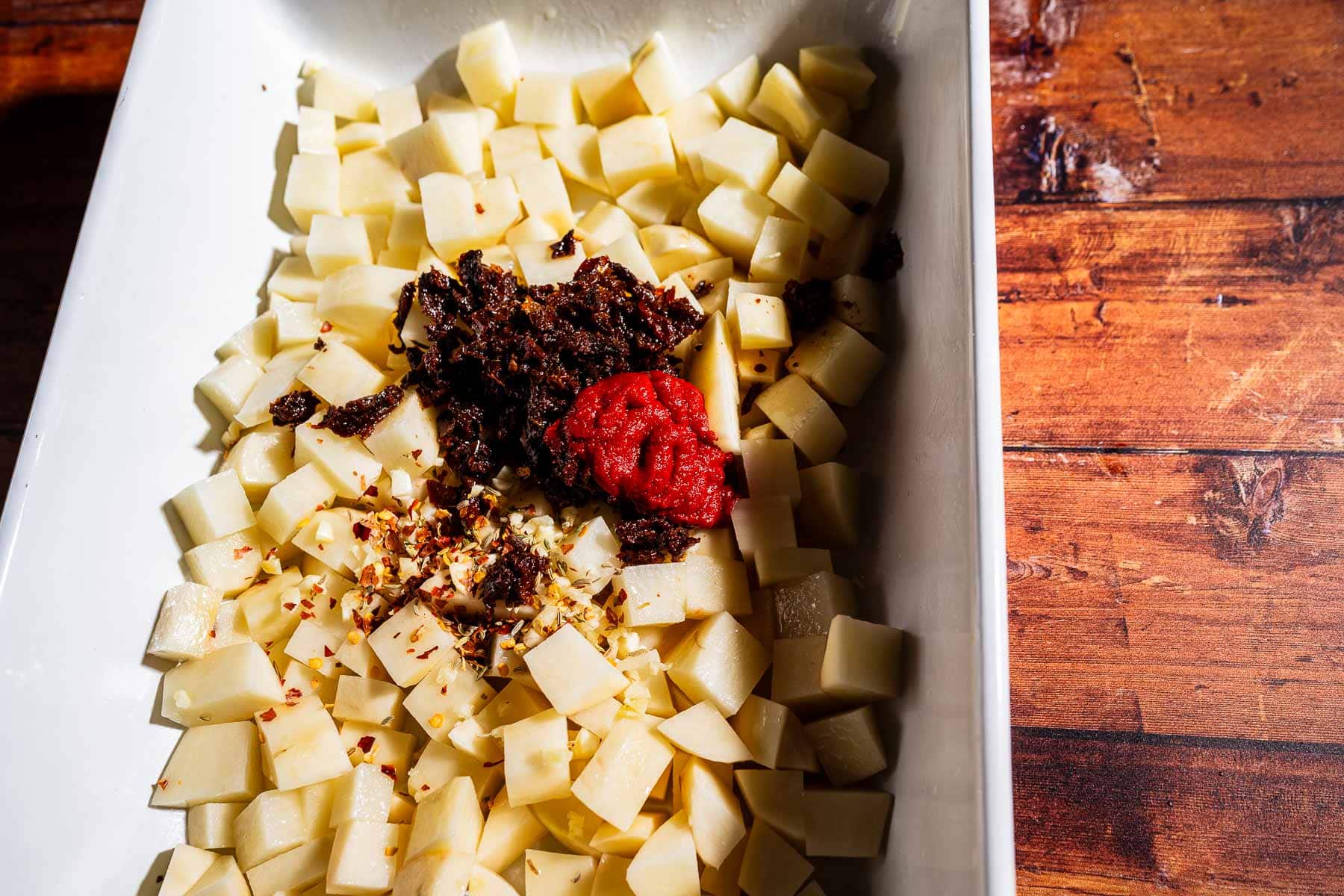Diced potatoes in a white dish topped with chopped sun-dried tomatoes, tomato paste, minced garlic, and red pepper flakes—a delicious spin on marry me potatoes—served on a wooden surface.