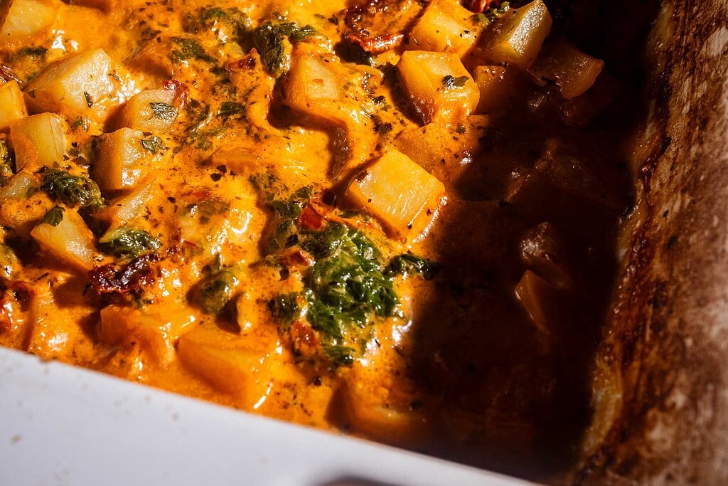 Close-up of a baked dish featuring marry me potatoes, leafy greens, and a creamy orange sauce, all nestled in a white baking dish.