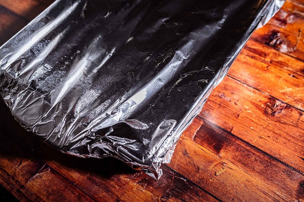 A rectangular baking dish covered with aluminum foil sits on a wooden surface, ready to reveal a batch of delicious marry me potatoes.