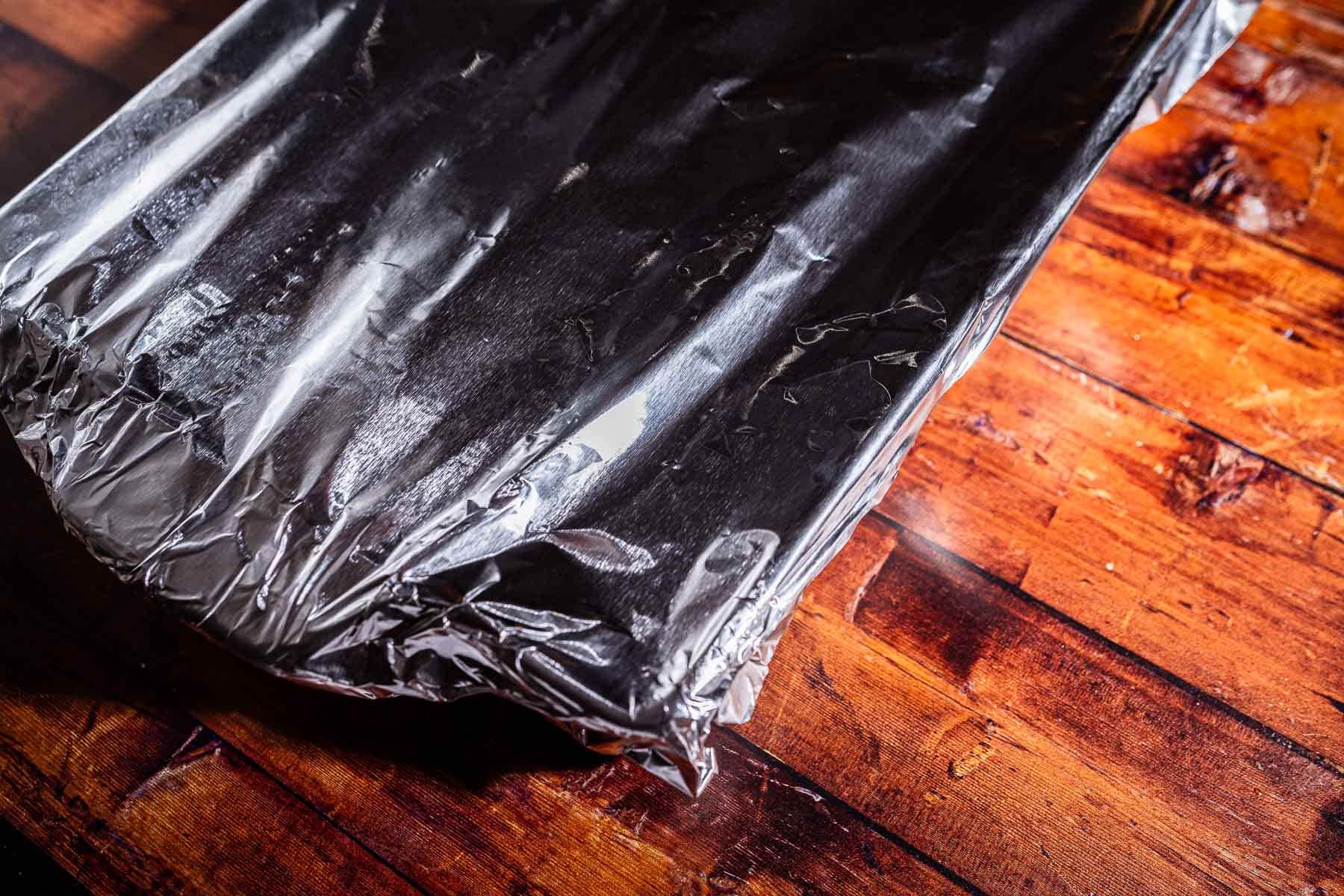 A rectangular baking dish covered with aluminum foil sits on a wooden surface, ready to reveal a batch of delicious marry me potatoes.