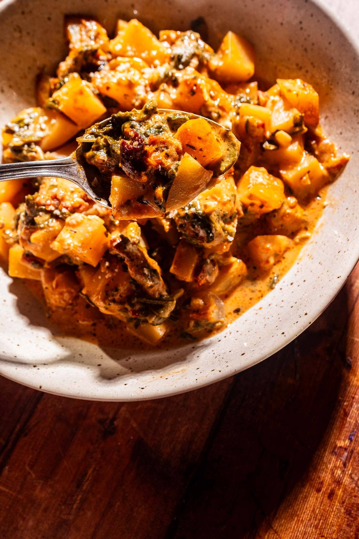 A bowl of creamy vegetable stew with diced marry me potatoes and greens, with a spoon lifting a portion from the bowl, on a wooden surface.
