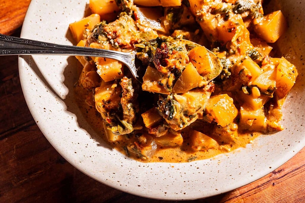 A close-up of a bowl filled with creamy stew, featuring marry me potatoes alongside cubed root vegetables and leafy greens, with a metal spoon lifting a hearty portion.
