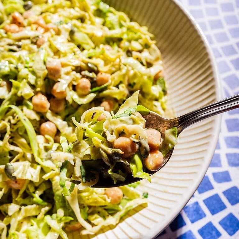 A close-up of a vibrant cabbage salad with chickpeas and seeds, with a spoonful being lifted over a blue and white tiled surface.