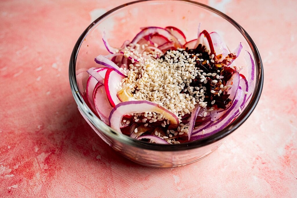 A glass bowl containing sliced red onions, sesame seeds, chili flakes, and sauce on a pink textured surface—perfect for adding to your favorite spicy radish recipe.
