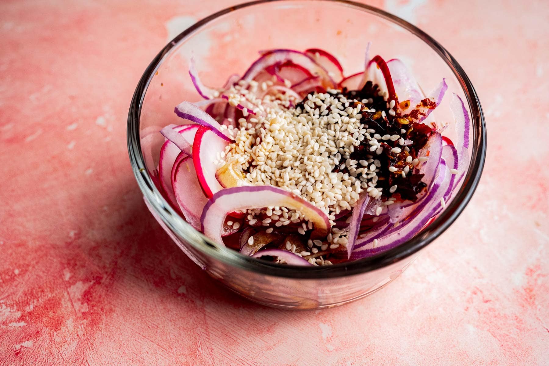 A glass bowl containing sliced red onions, sesame seeds, chili flakes, and sauce on a pink textured surface&mdash;perfect for adding to your favorite spicy radish recipe.