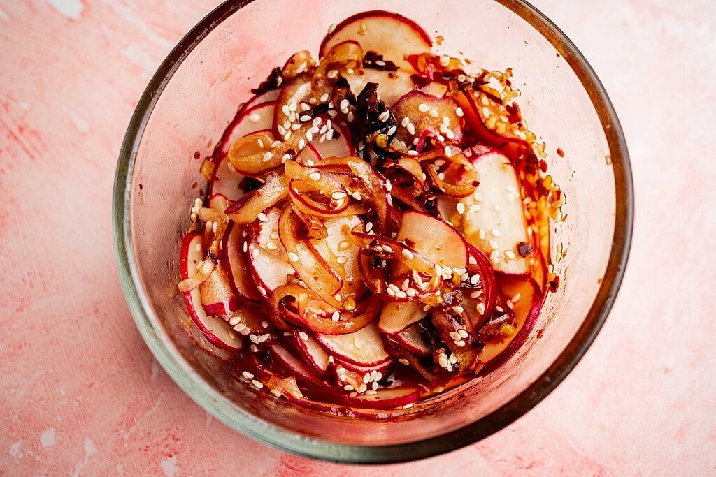 Thinly sliced radishes tossed with sesame seeds and seasoning create a vibrant spicy radish recipe, beautifully arranged in a glass bowl and photographed from above on a pink surface.