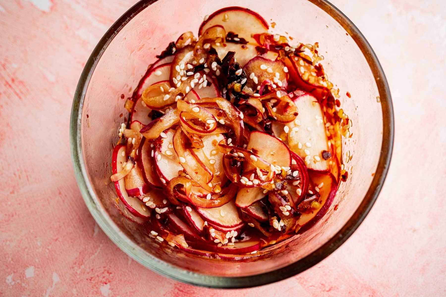 Thinly sliced radishes tossed with sesame seeds and seasoning create a vibrant spicy radish recipe, beautifully arranged in a glass bowl and photographed from above on a pink surface.
