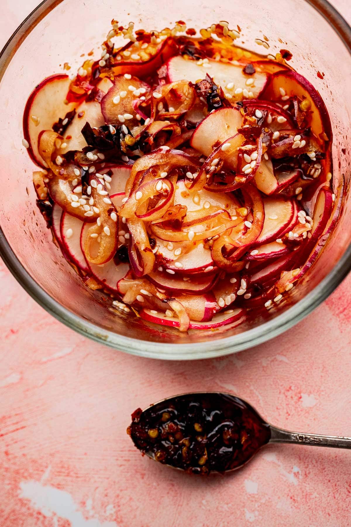 Glass bowl with sliced radishes and onions tossed in sesame seeds and chili oil; a spoon with chili oil rests on the pink surface nearby, making this a vibrant spicy radish recipe.